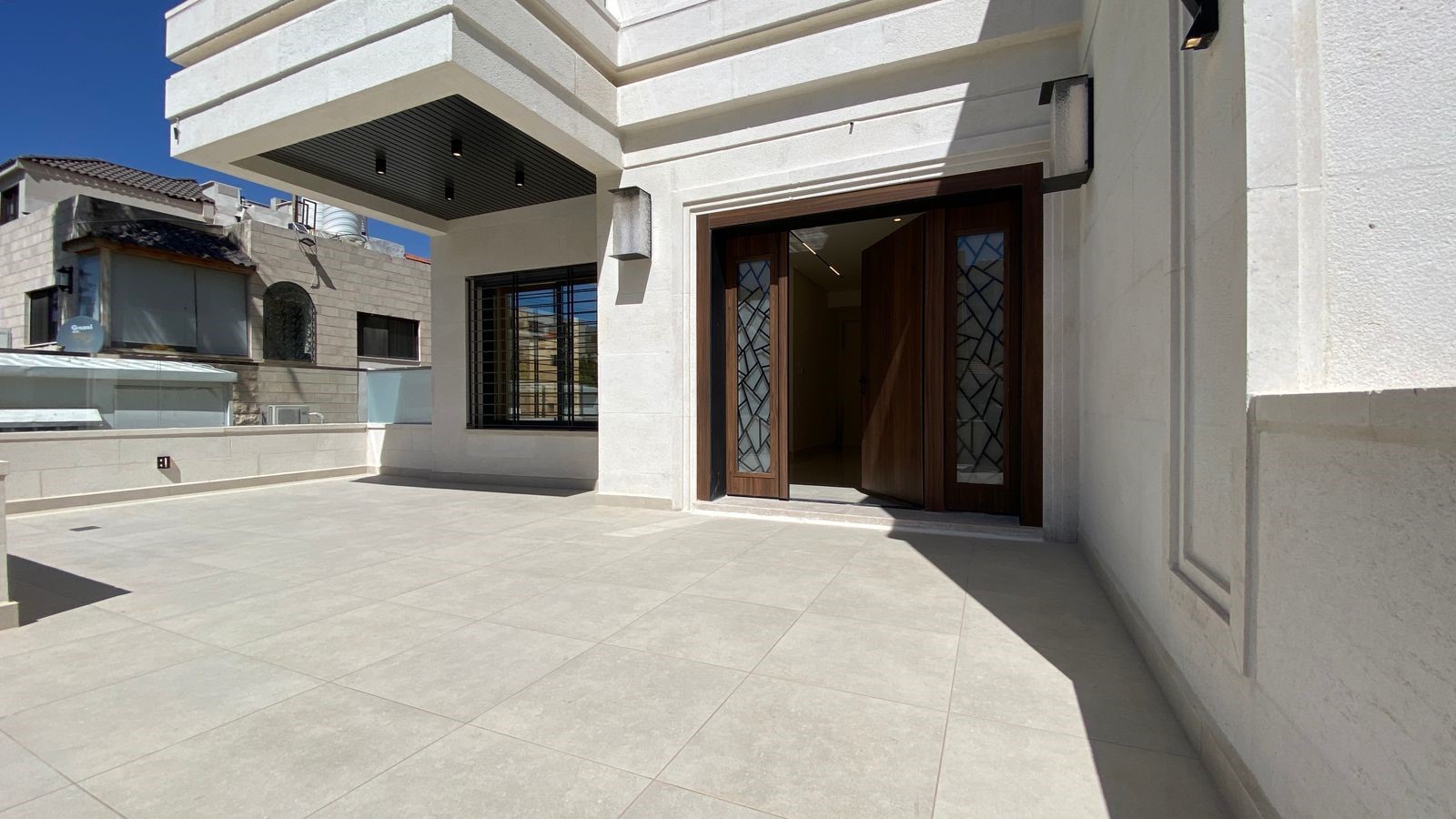 e6cb3902-5751-4094-bb23-9ca4be916834 Open double wooden doors with decorative glass panels reveal a lit interior, on a large sunlit tiled terrace of a modern white building.