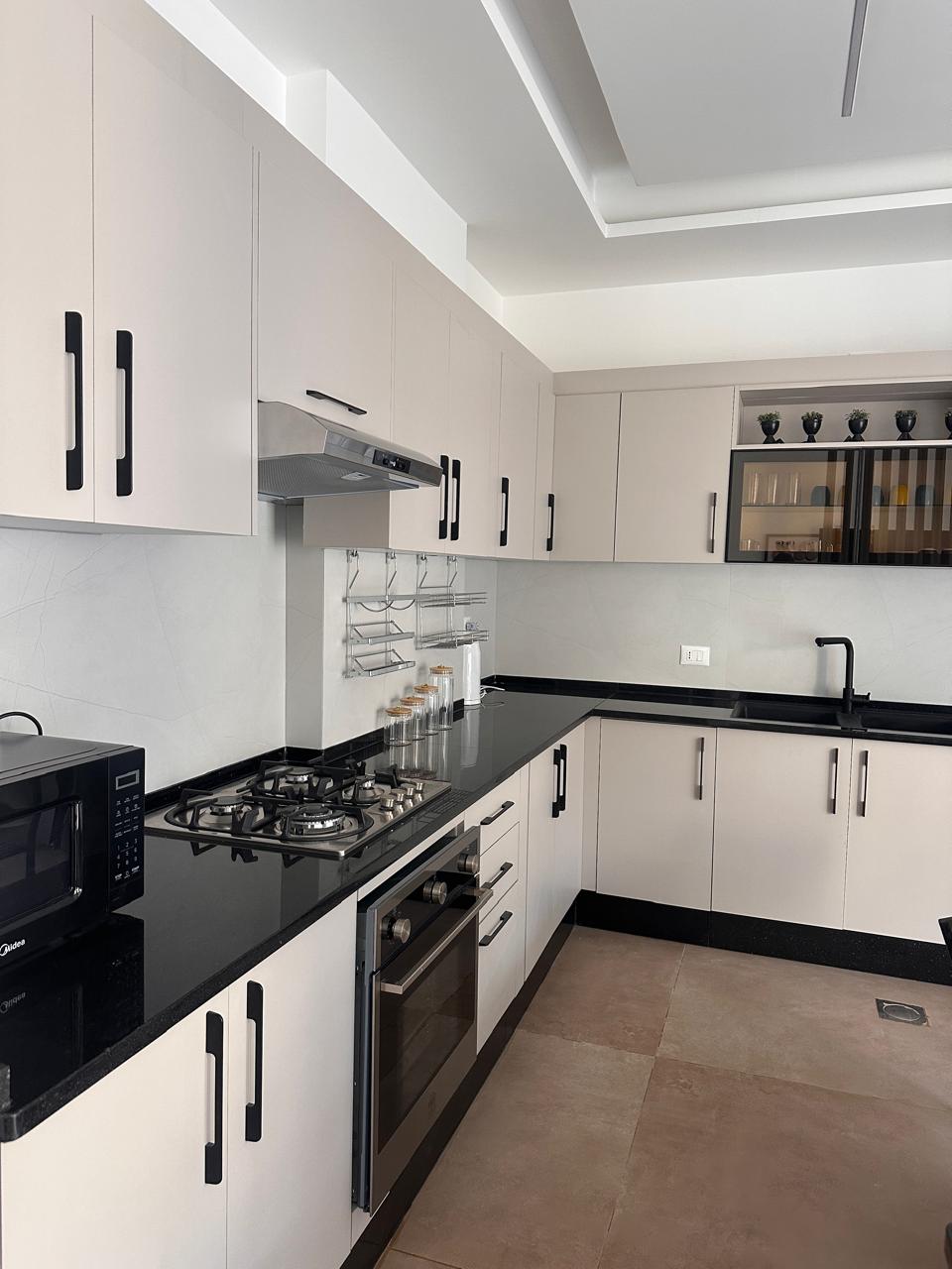 e7fe71df-76ee-44d3-b8bc-e598fd3523bd Modern kitchen with white cabinets, black countertops, and stainless steel stove and oven layout along an L-shaped wall.