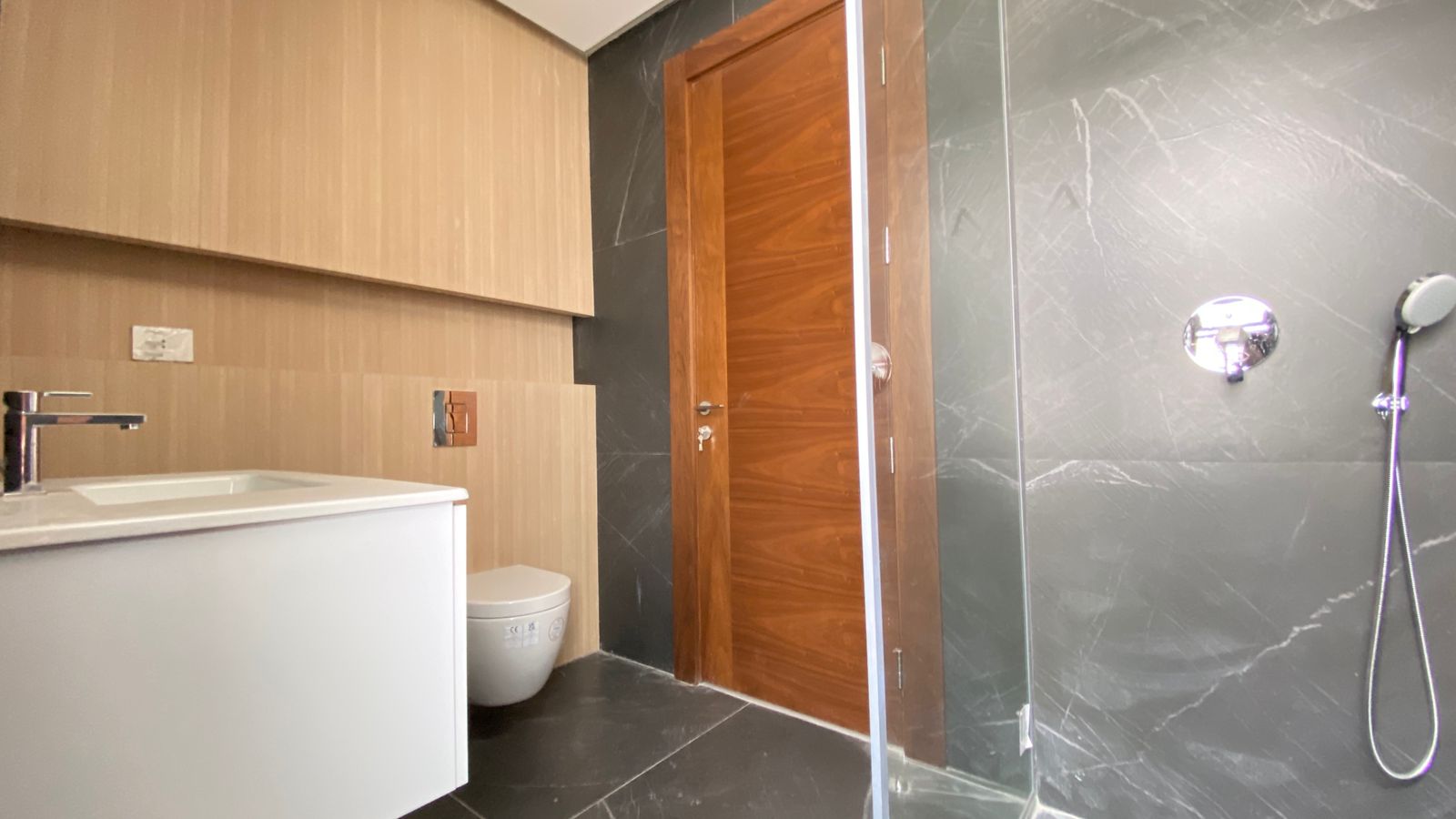e9303c3d-417b-4bee-b2c4-e1dd5f7459cc Modern bathroom with beige wood-paneled walls, white vanity, wall-hung toilet, wooden door, and a glass shower with handheld shower.