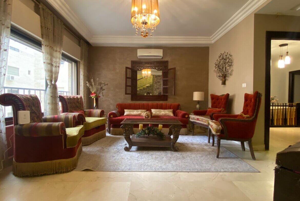 e9e5933d-46d4-47c4-b99a-9bf7b3ea990a Elegant living room with red velvet sofa and armchairs, ornate chandelier, and a wood coffee table on a light rug on marble floor.