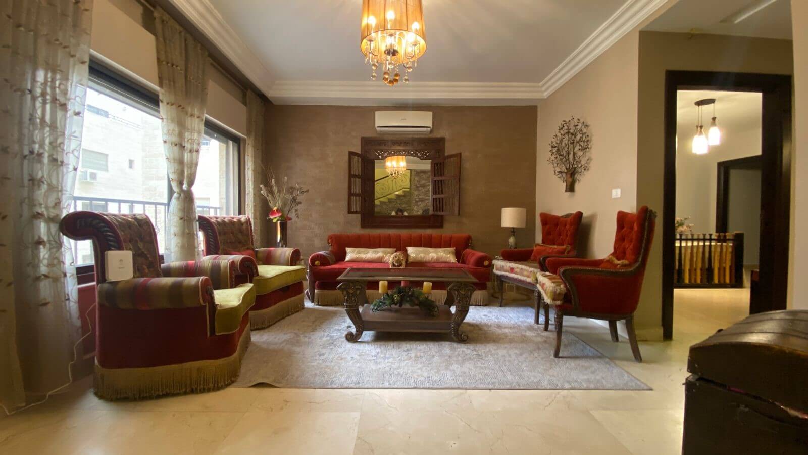 e9e5933d-46d4-47c4-b99a-9bf7b3ea990a Elegant living room with red velvet sofa and armchairs, ornate chandelier, and a wood coffee table on a light rug on marble floor.