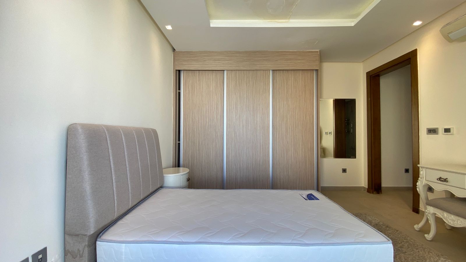ea5f830d-4e2c-423a-a0bc-0af1c893fbc4 Modern bedroom with a padded headboard bed, a large wooden sliding-door wardrobe, and a white dresser visible in a bright, minimalist room.