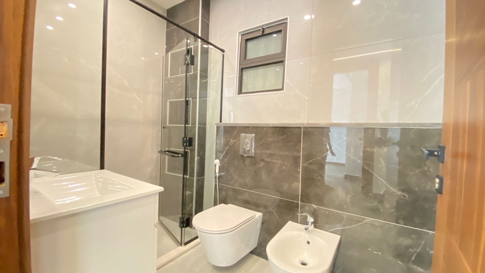 ea8a16d3-5654-44ba-a789-a33080dbc03d 1 Modern bathroom with white vanity, wall-mounted toilet and bidet, glass shower enclosure, and gray marble tiles.