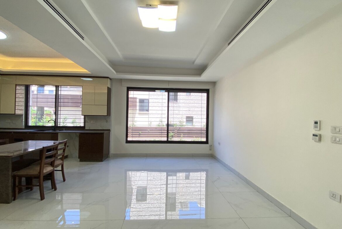 Open-plan kitchen and dining area with a marble-top table, wooden chairs, white glossy floor, and a large window bringing in daylight.