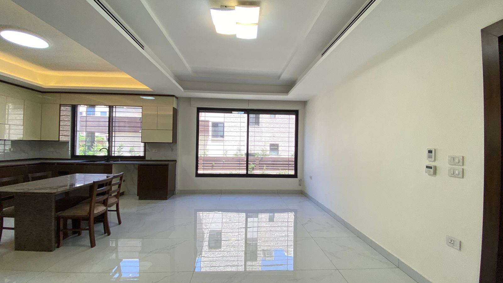 eaf7f558-f6bc-428d-9e73-f240273028fa Open-plan kitchen and dining area with a marble-top table, wooden chairs, white glossy floor, and a large window bringing in daylight.