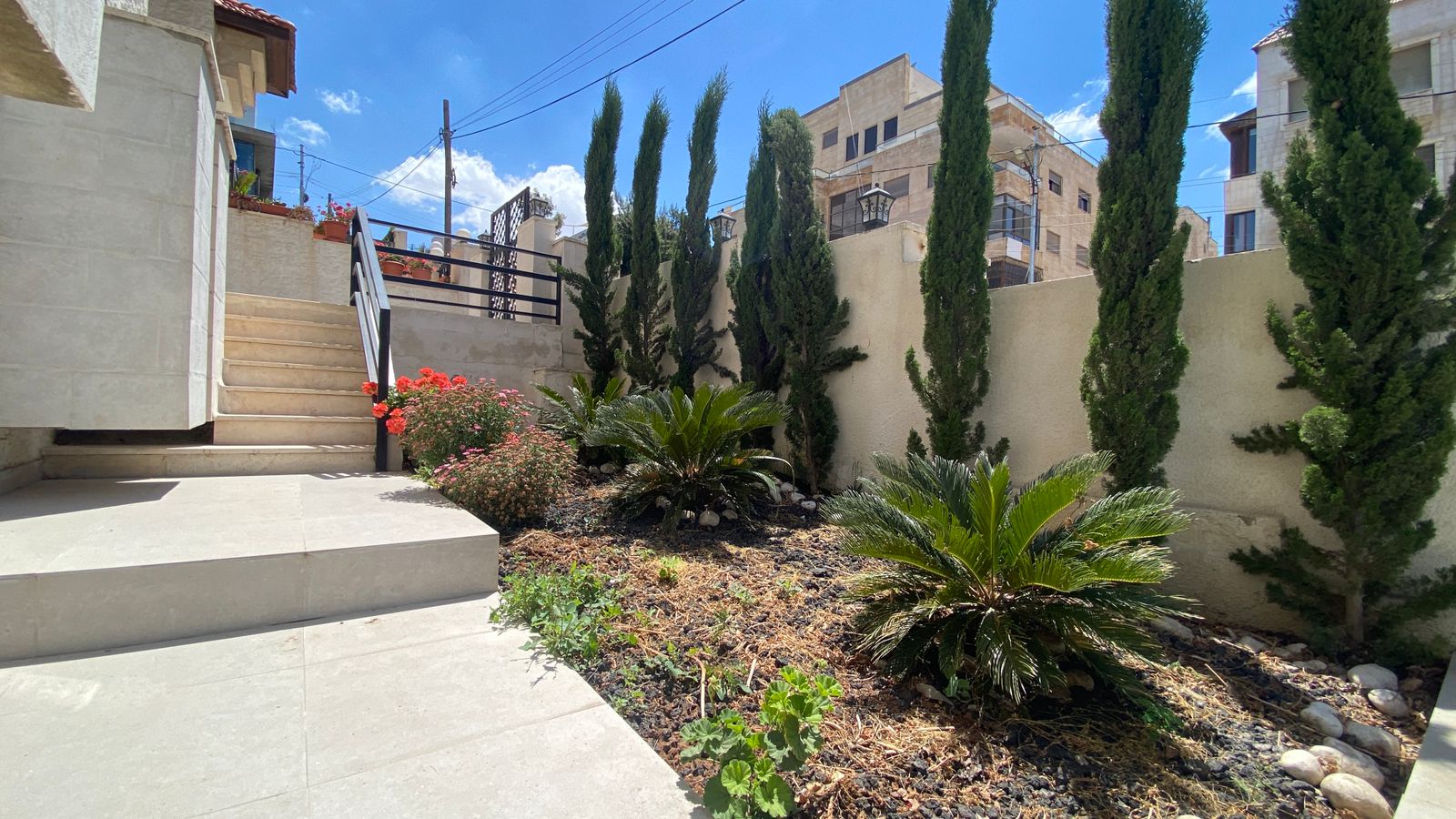 eb690f74-fd23-4007-945a-03d8fa82bc69 Stone patio with steps up to a railing, beige walls, and a garden bed with palms and tall cypress trees against a blue sky in a sunny courtyard.