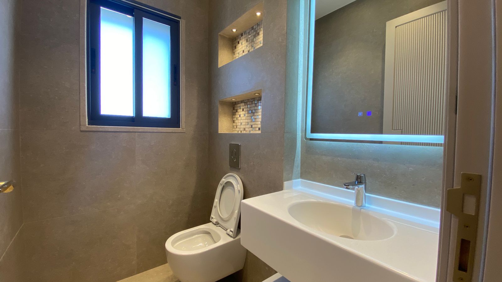 eb8ecaa6-df5e-478d-9836-d7900470b537 Modern bathroom with wall-to-wall beige tiles, a frosted window, and a white wall-mounted sink with backlit mirror lighting the room behind.
