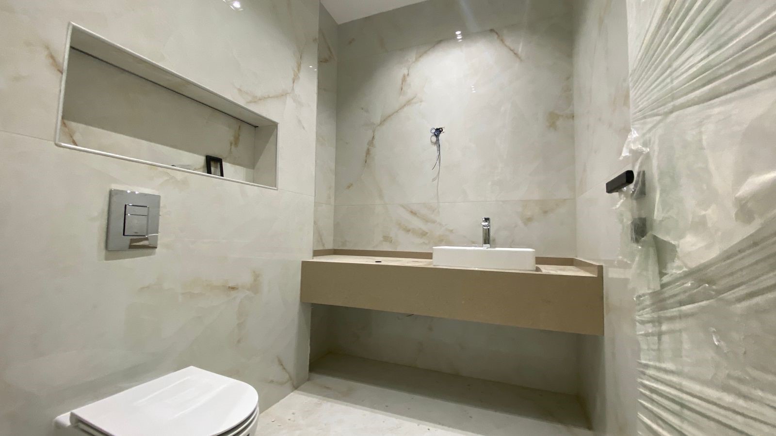 ec0b8670-b922-4e93-92a7-dd9f41b054a5 Modern bathroom under construction with marble walls, recessed shelf, wall-mounted toilet, and beige floating vanity with white sink.