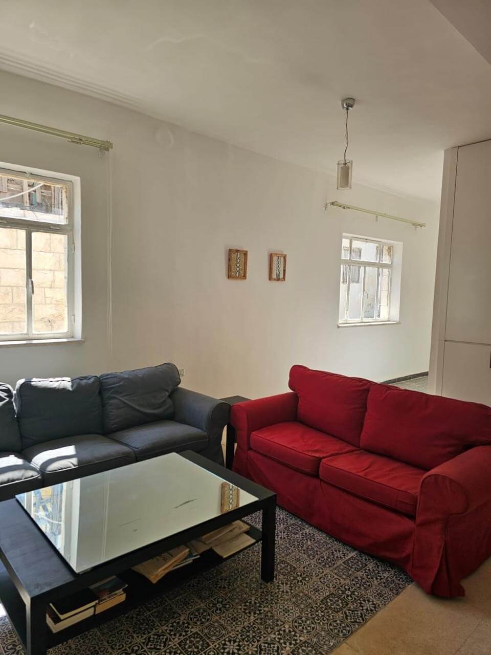 ec7aa2e1-d196-40a7-95a6-2df305eb2586 Living room with a gray sofa and a red sofa around a glass-topped coffee table on a patterned rug. Two windows let in light and small wall decorations hang between them.