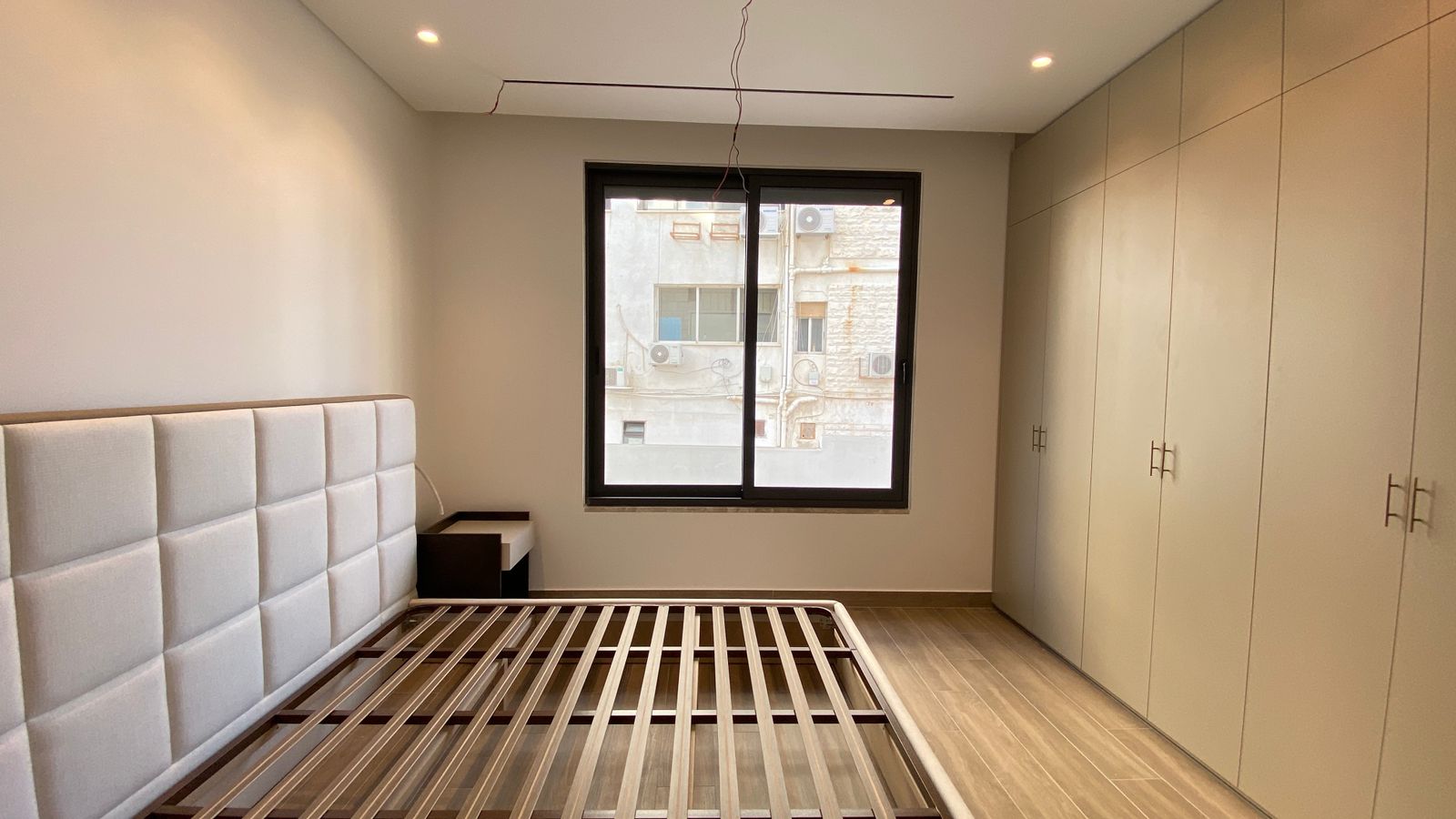 ec82c642-bcae-459b-b953-8e889cb6e2a1 Minimalist bedroom with a padded headboard bed frame, a small nightstand, and tall built-in wardrobes along the right wall. A large window faces an urban exterior.