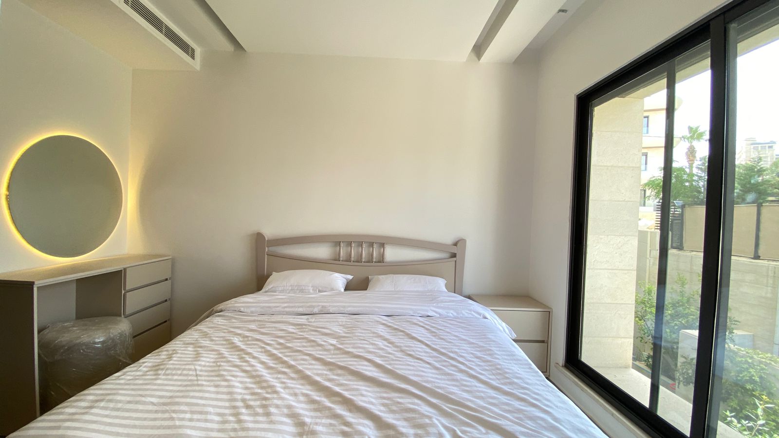 ed540777-db67-422e-b3cc-f393adaab02e Minimalist bedroom with a bed centered on the wall, a circular backlit mirror above a dresser on the left, and a large window on the right.