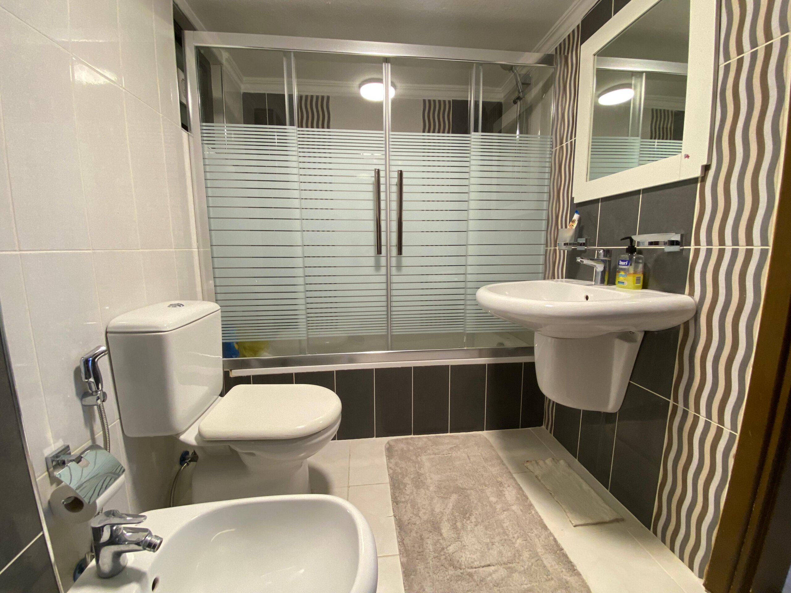 edf23718-9459-419e-b9dc-e085c881d2b8 Modern bathroom with a glass-paneled shower enclosure featuring frosted horizontal stripes, and a white toilet and pedestal sink with chrome fixtures nearby.