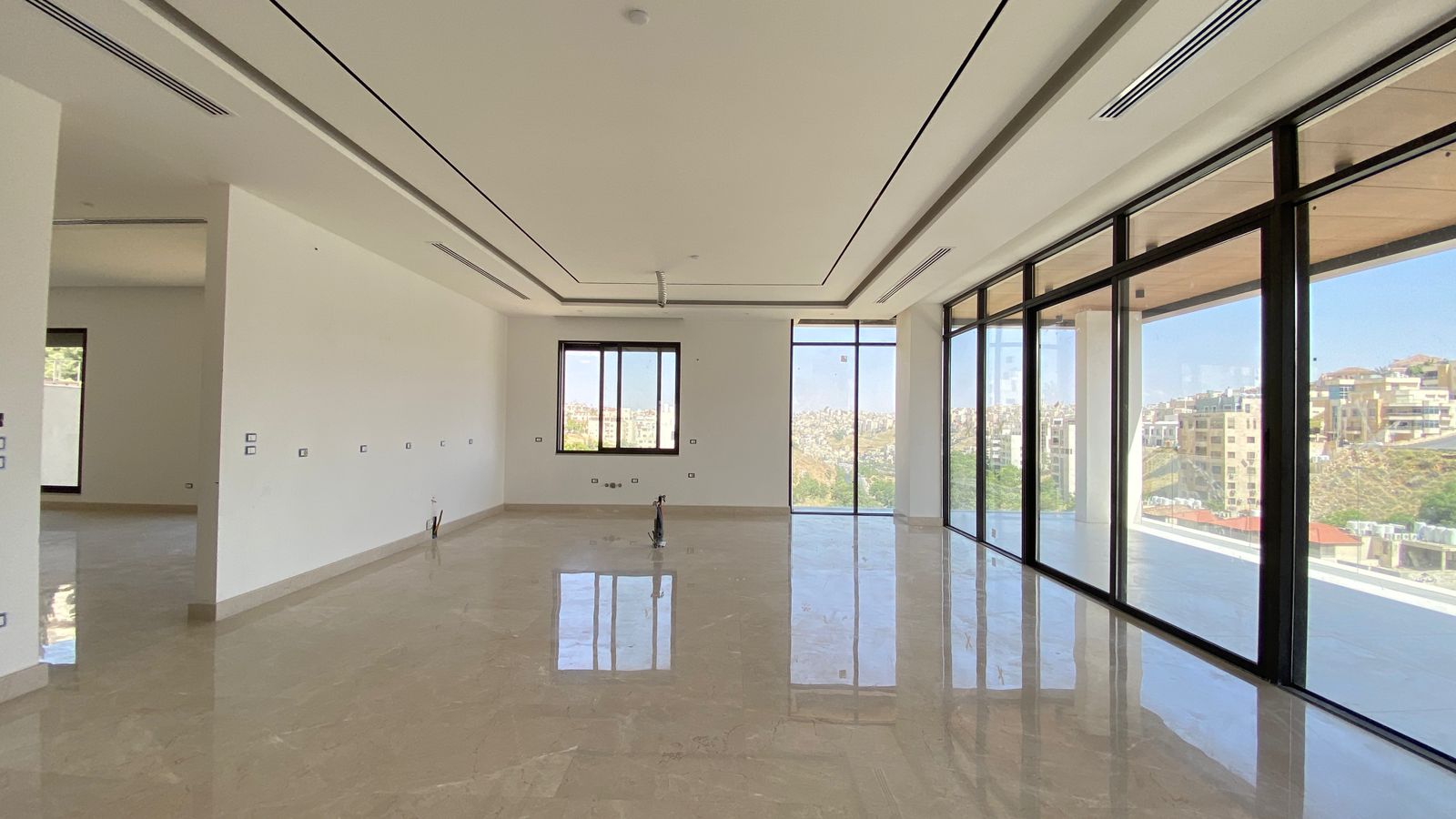 ee03a619-b410-40d5-9717-39a062a914d7 Spacious empty living area with polished marble floor and floor-to-ceiling glass windows overlooking a cityscape.