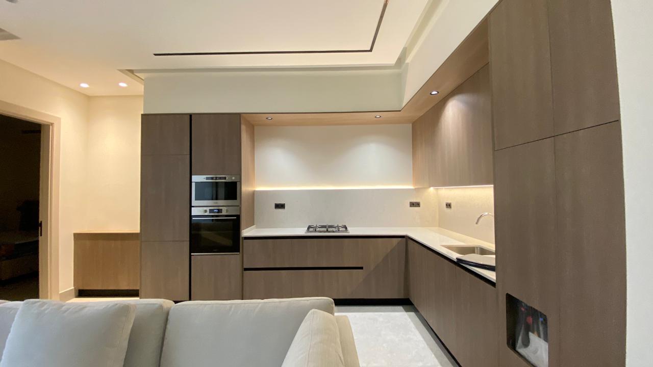 Contemporary L-shaped kitchen with dark wood cabinetry, built-in oven/microwave, white countertops, and under-cabinet lighting.
