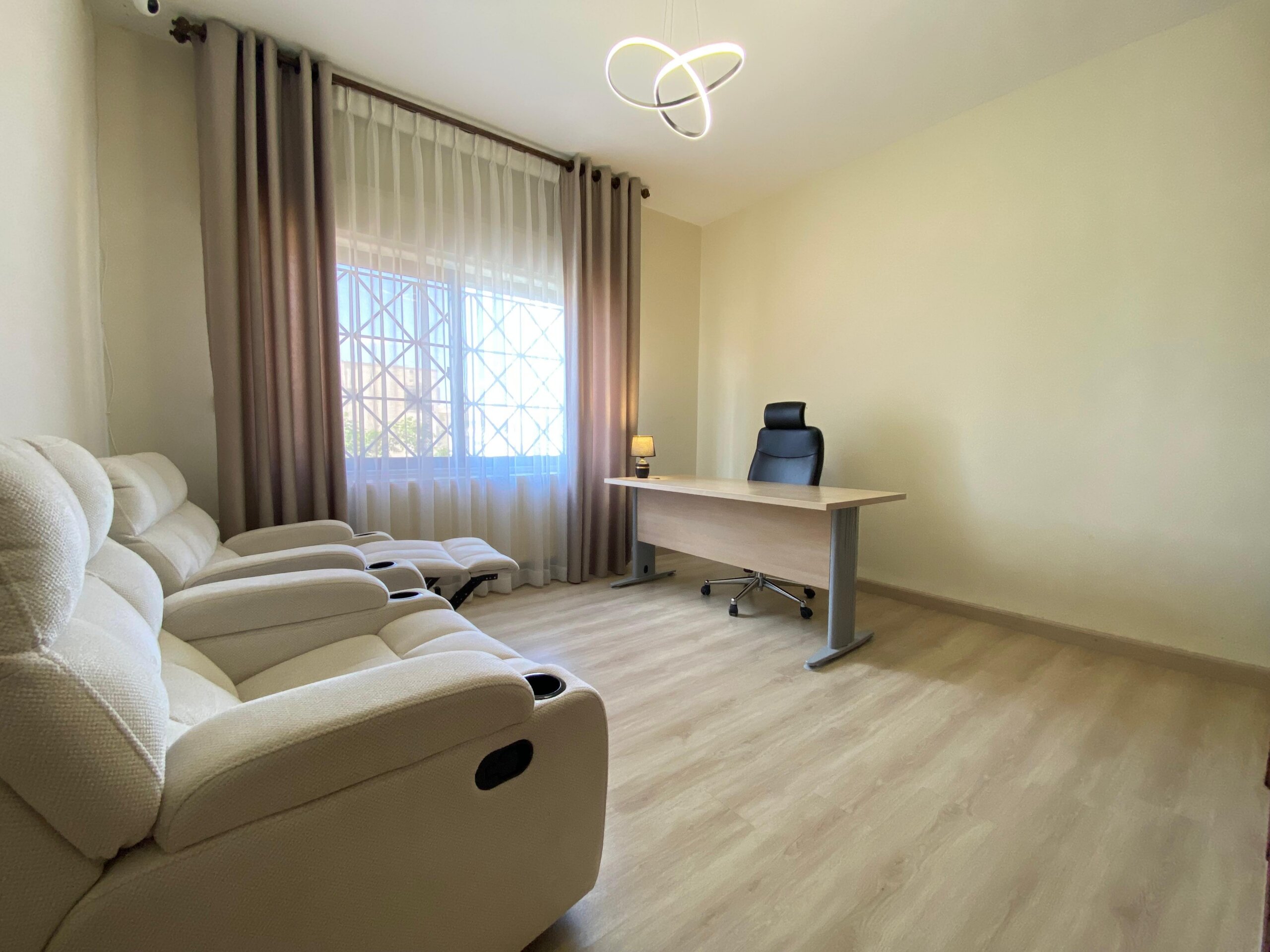 eea0a1a6-3e65-4a33-b737-c11da1db3b16 Living room with a beige recliner sofa, a large window with sheer and draped curtains, and a wooden desk with a black ergonomic chair nearby.