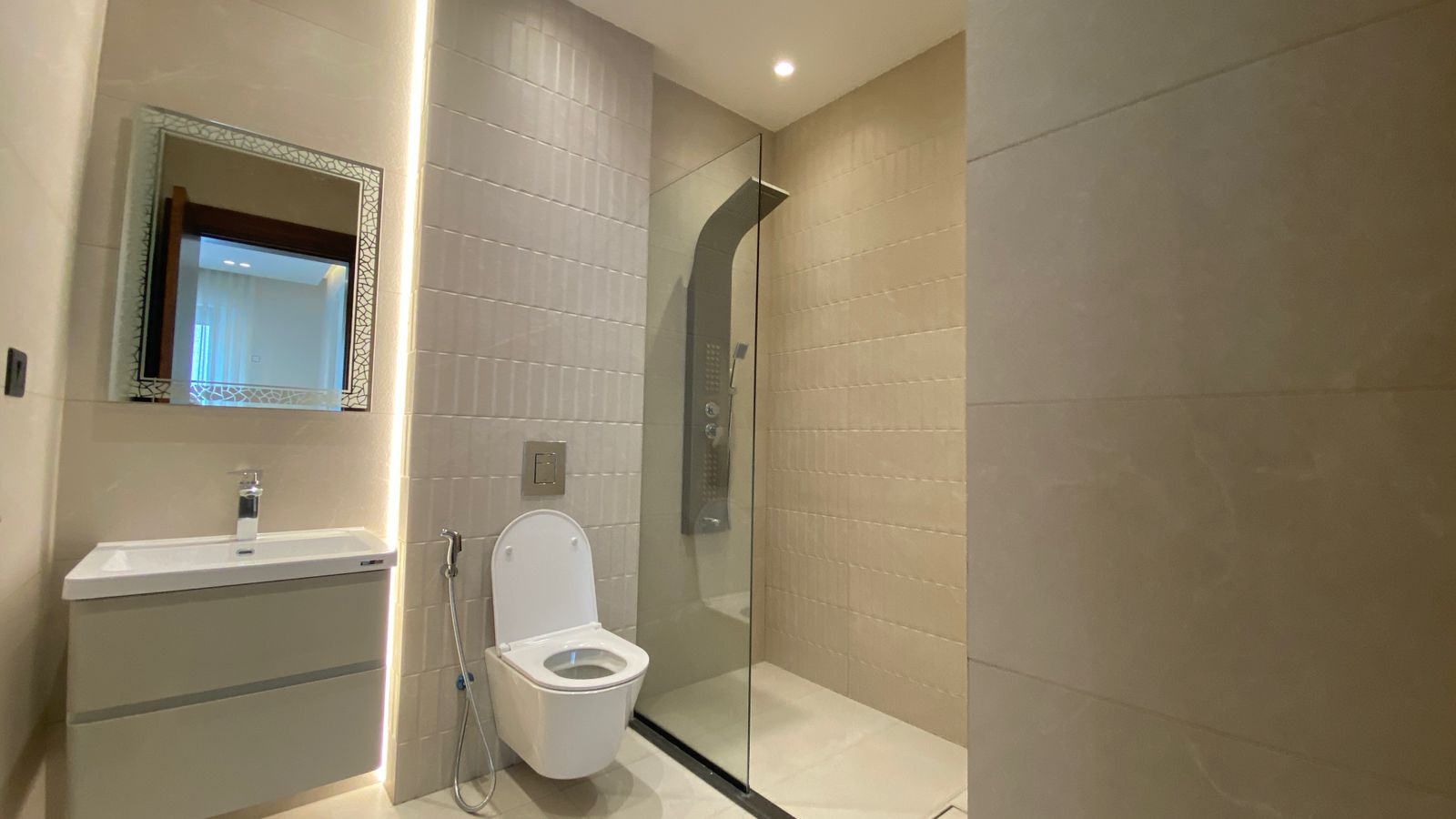 ef3ff390-4d3a-4ed7-b2d8-8c3fbe39a1cd Modern bathroom with wall-mounted vanity, backlit decorative mirror, wall-hung toilet, handheld bidet spray, and a glass-enclosed shower.