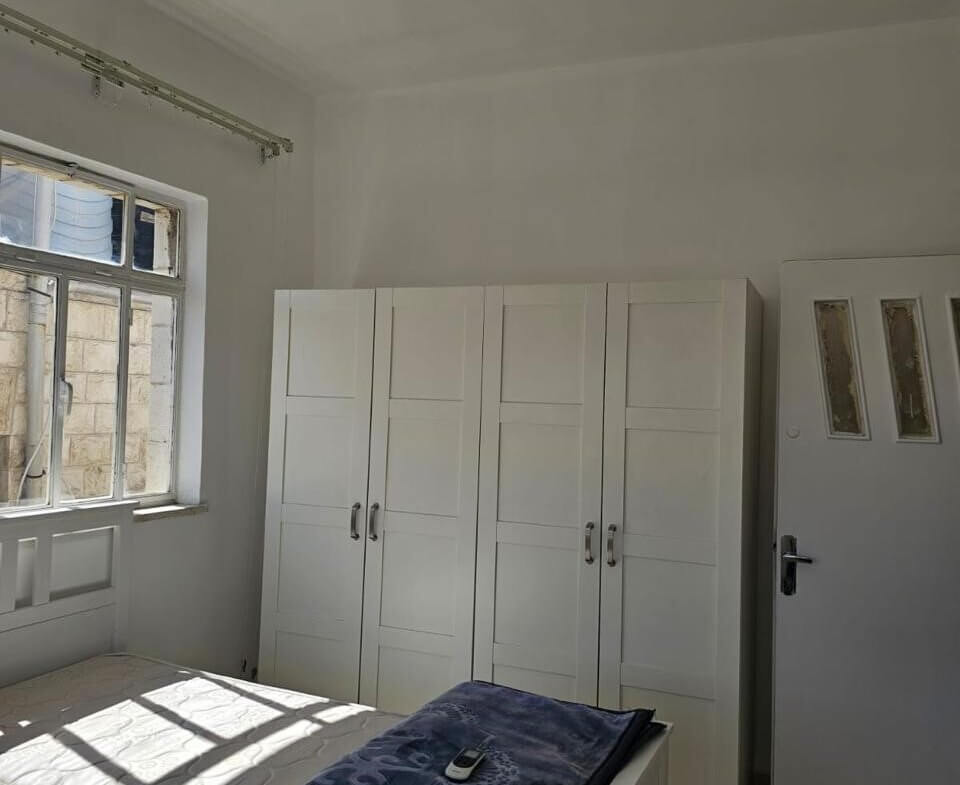 ef4a47cf-b7a2-4fd3-89cc-4e392c7d13e7 Bright white bedroom with a bed, a blue blanket and remote on top, white wardrobes along the wall, and a window letting in sunlight.