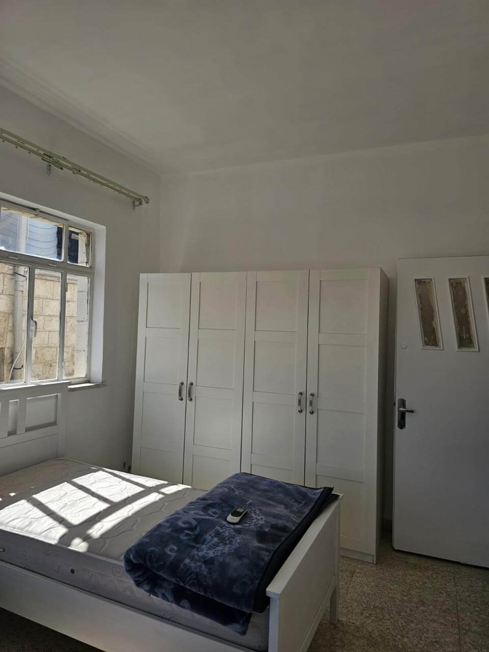 ef4a47cf-b7a2-4fd3-89cc-4e392c7d13e7 Bright white bedroom with a bed, a blue blanket and remote on top, white wardrobes along the wall, and a window letting in sunlight.