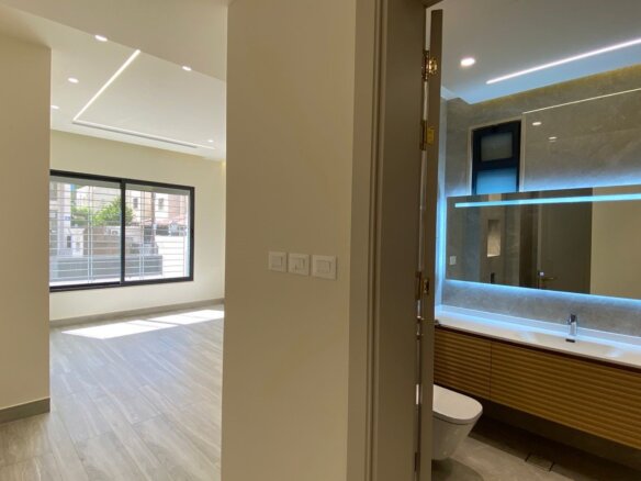 Modern open-plan interior: bright living area with large window, light wood floors, and recessed ceiling lights; adjacent bathroom visible through doorway with illuminated backlit mirror and wooden vanity.