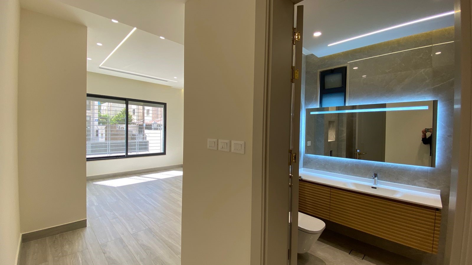 efb589b8-a46b-4b5f-bc82-fa008212c478 Modern open-plan interior: bright living area with large window, light wood floors, and recessed ceiling lights; adjacent bathroom visible through doorway with illuminated backlit mirror and wooden vanity.