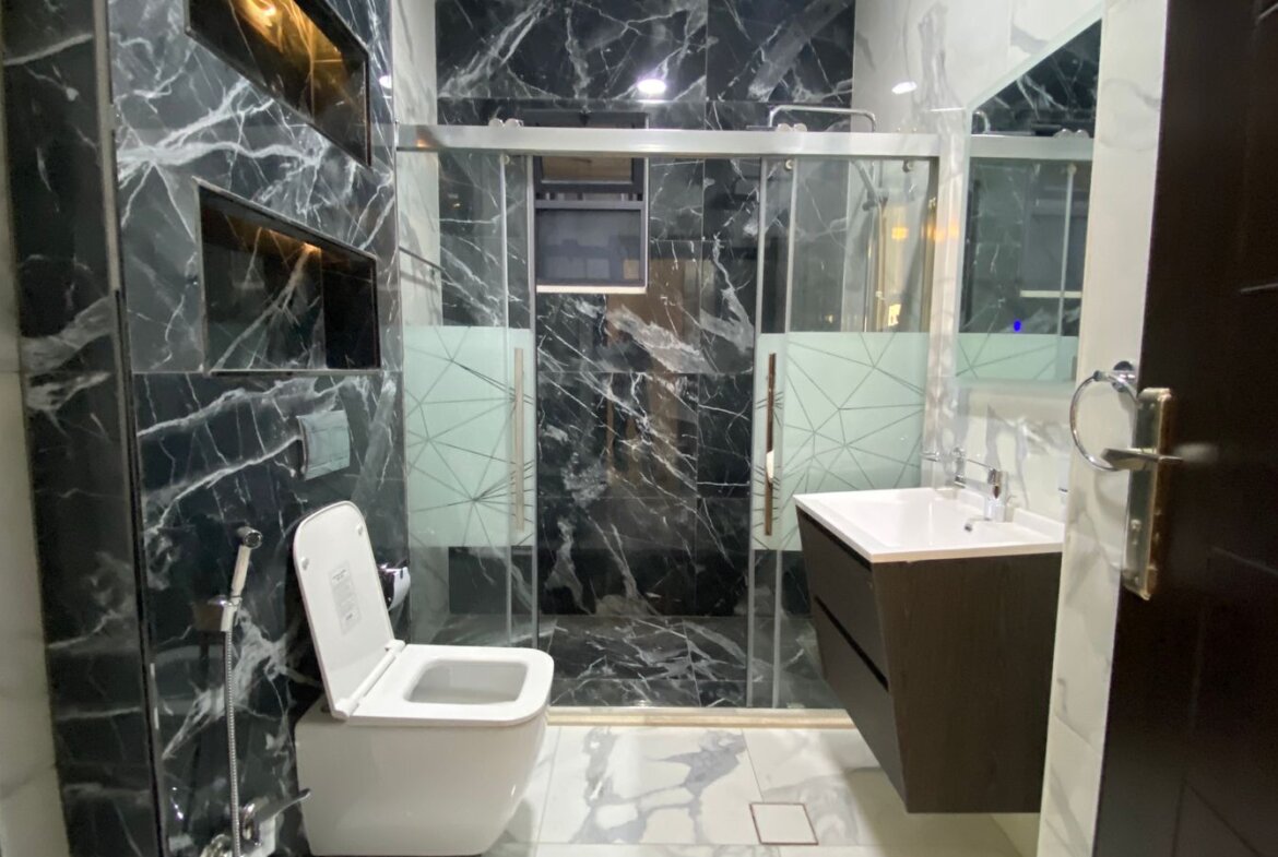 Modern bathroom with black marble walls, white toilet, glass-framed shower, and dark wood vanity with white sink.