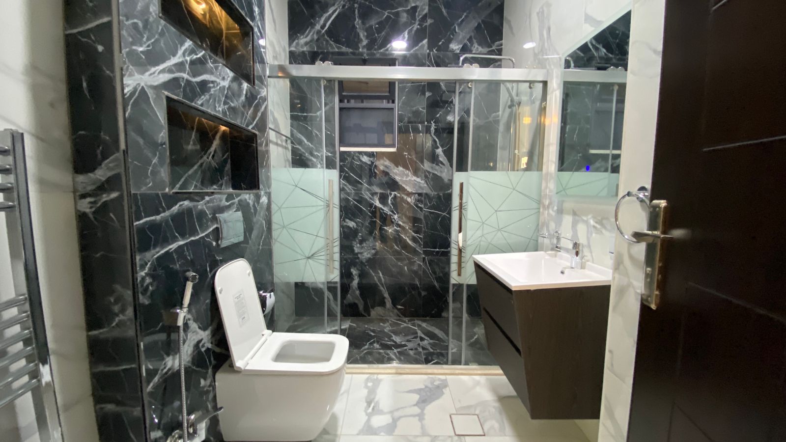Modern bathroom with black marble walls, white toilet, glass-framed shower, and dark wood vanity with white sink.