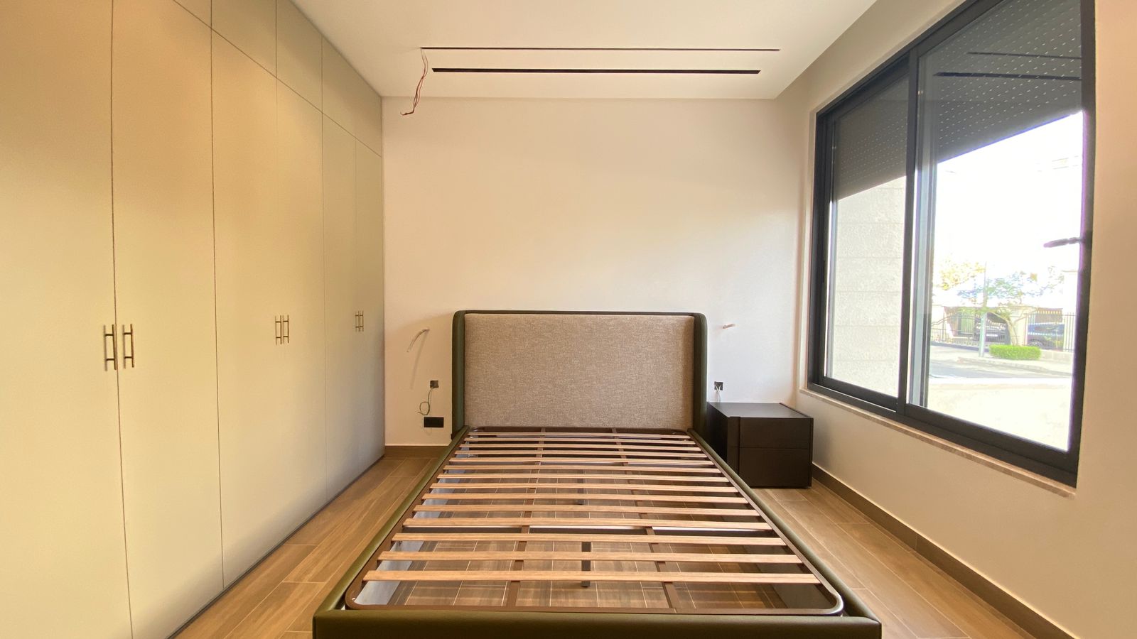 f06fec93-4154-4d14-b2ae-1a2e7b6b90c7 Modern bedroom with an empty bed frame and gray upholstered headboard, beige built-in wardrobes along the left wall, and a large window on the right showing an outdoor scene.