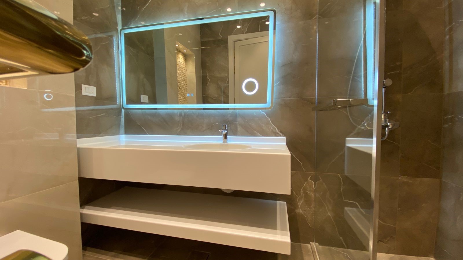 f0e53c1c-73b9-4690-b0cd-cc34cf9bbad7 Modern bathroom with a backlit rectangular mirror above a floating white sink and an open shelf, marble walls, and a glass shower enclosure to the right