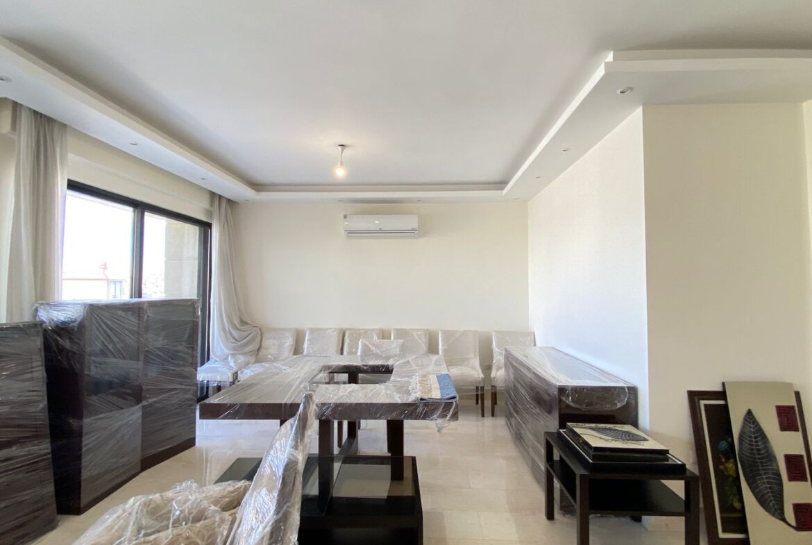 Living room with white walls and a modern ceiling, large window with sheer curtains, and sofas and tables covered in plastic wrap.