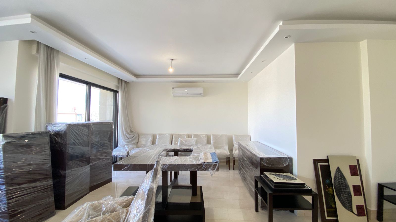 f15aecc0-7ee9-49a3-b7d2-321b1d3dcd5a Living room with white walls and a modern ceiling, large window with sheer curtains, and sofas and tables covered in plastic wrap.