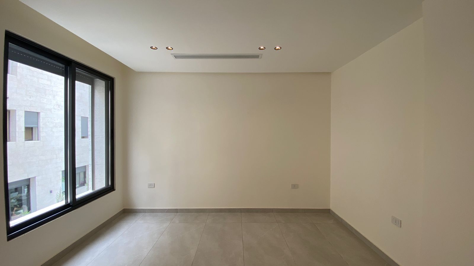 f1c02140-9893-4aac-878f-7ca6ec20b79d Empty, unfurnished room with a large black-framed window along the left wall, beige walls, and light gray tiled floor, plus recessed ceiling lights and a central air vent.