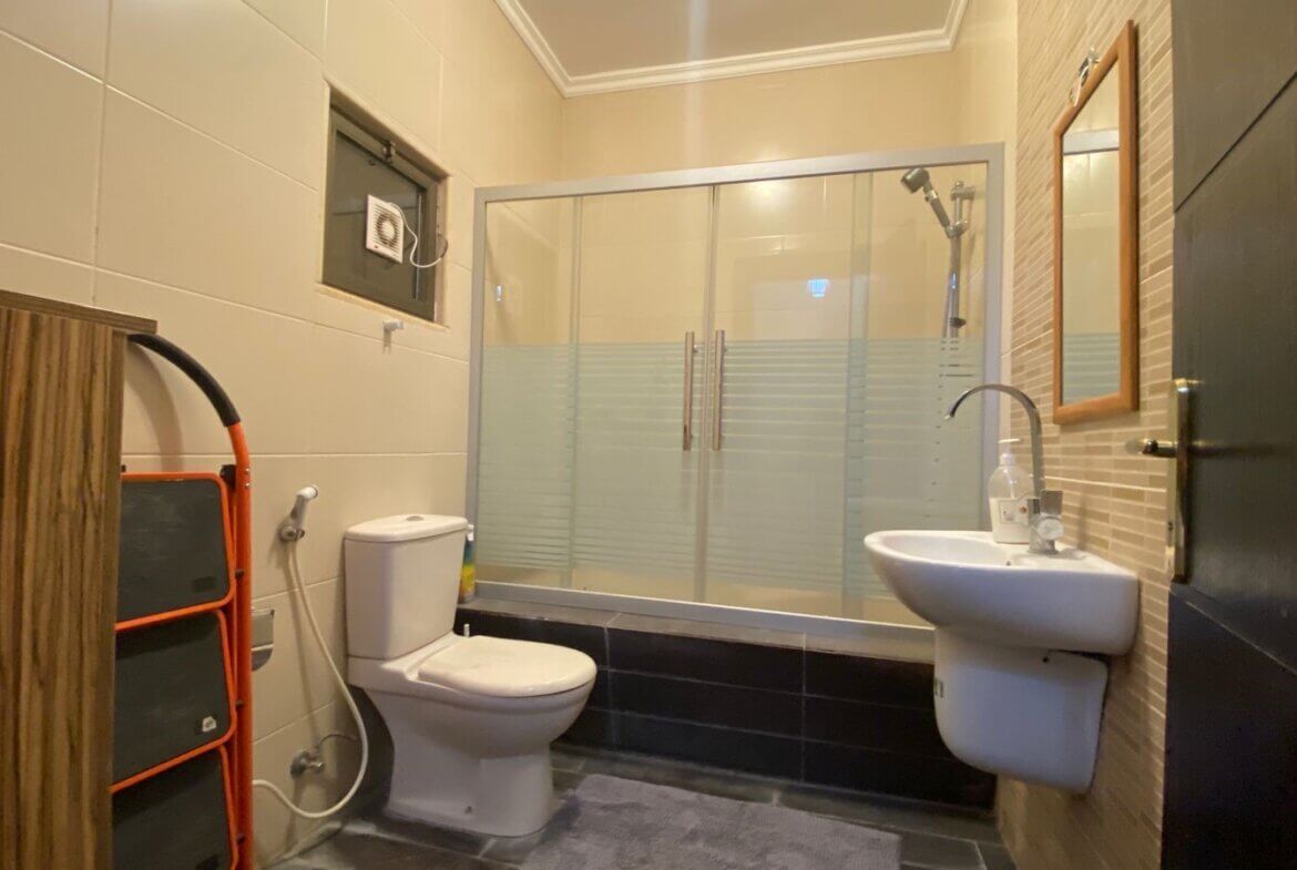 f24ef391-1e37-4f50-aa32-d33b48555312 Bathroom with beige tiled walls, glass-framed shower enclosure, toilet, and a wall-mounted sink.