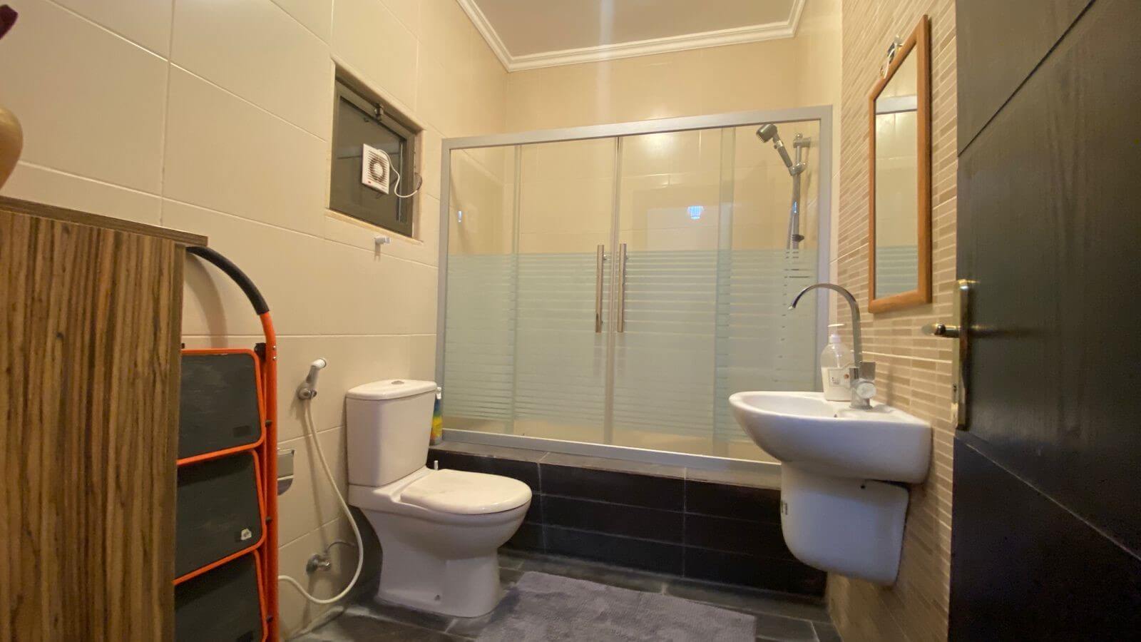 f24ef391-1e37-4f50-aa32-d33b48555312 Bathroom with beige tiled walls, glass-framed shower enclosure, toilet, and a wall-mounted sink.