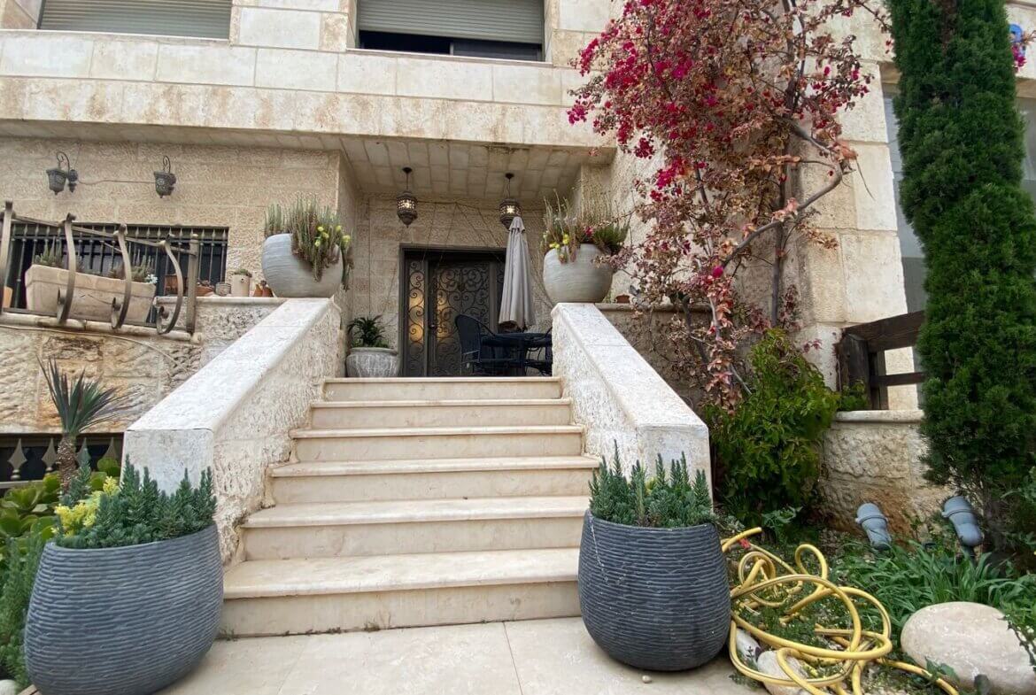 Stone staircase up to a wrought-iron door on a beige stone house, with large round planters and a coiled yellow hose nearby.