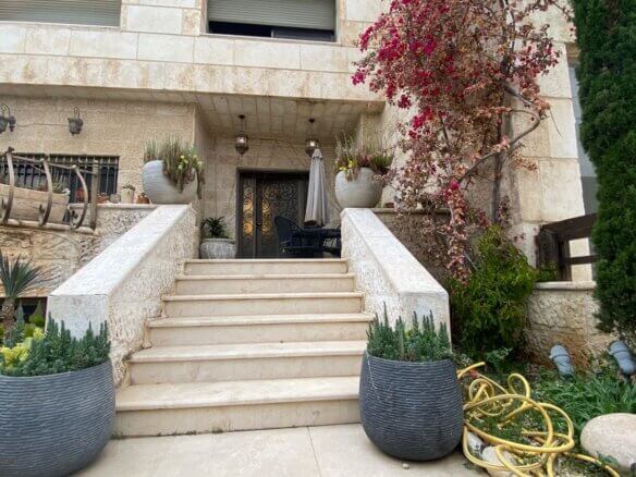 Stone staircase up to a wrought-iron door on a beige stone house, with large round planters and a coiled yellow hose nearby.