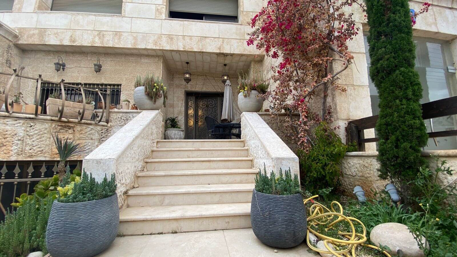f3112ed7-6c9d-40fd-824b-75300a5571d9 Stone staircase up to a wrought-iron door on a beige stone house, with large round planters and a coiled yellow hose nearby.