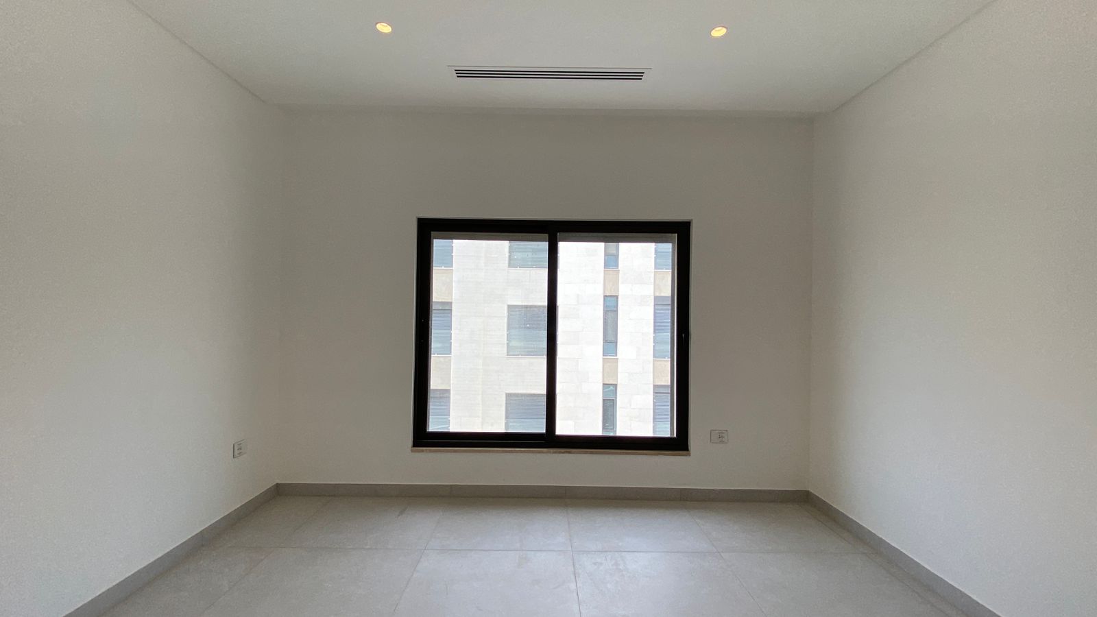 f3bf5dd3-8dc0-4319-9ab2-176c7468dd4a Empty, unfurnished room with white walls, gray tile floor, and a large central window showing a beige building outside; ceiling lights on.