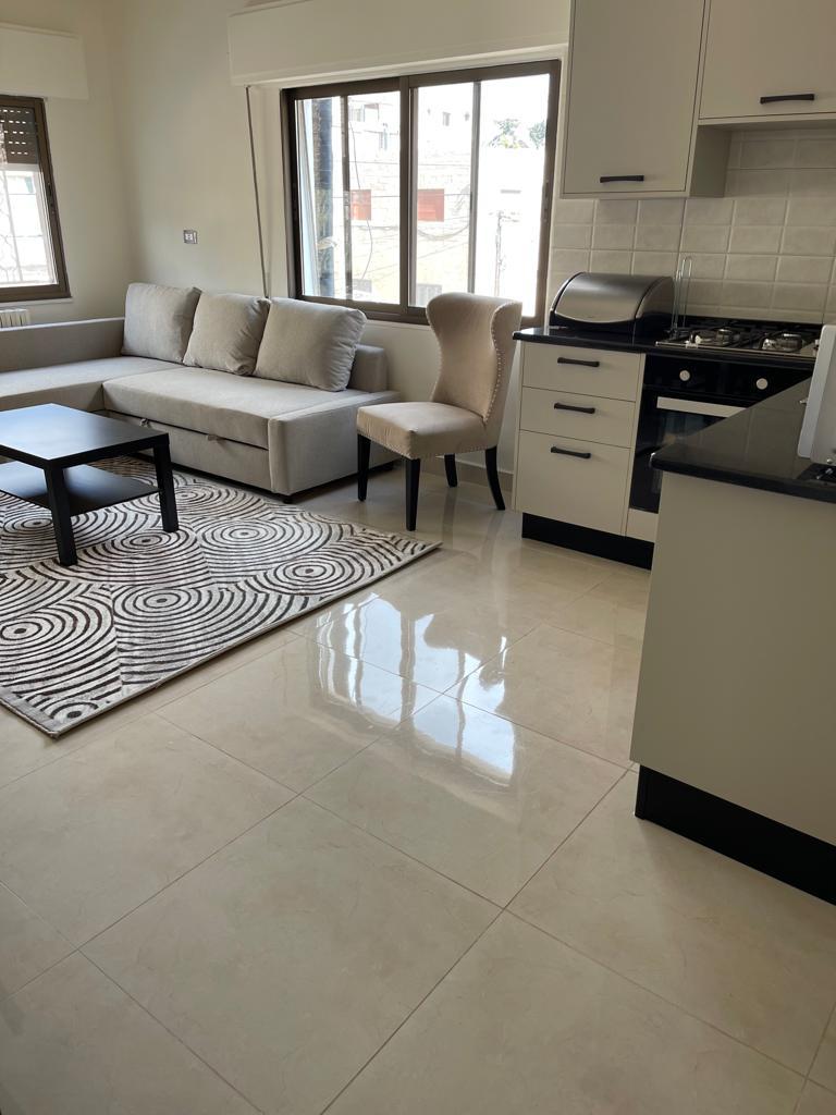 f3fb159f-f36f-46e2-bef5-2f20e07b2100 Open-plan living area with a gray sectional sofa, patterned rug, and black coffee table next to a windowed kitchen.