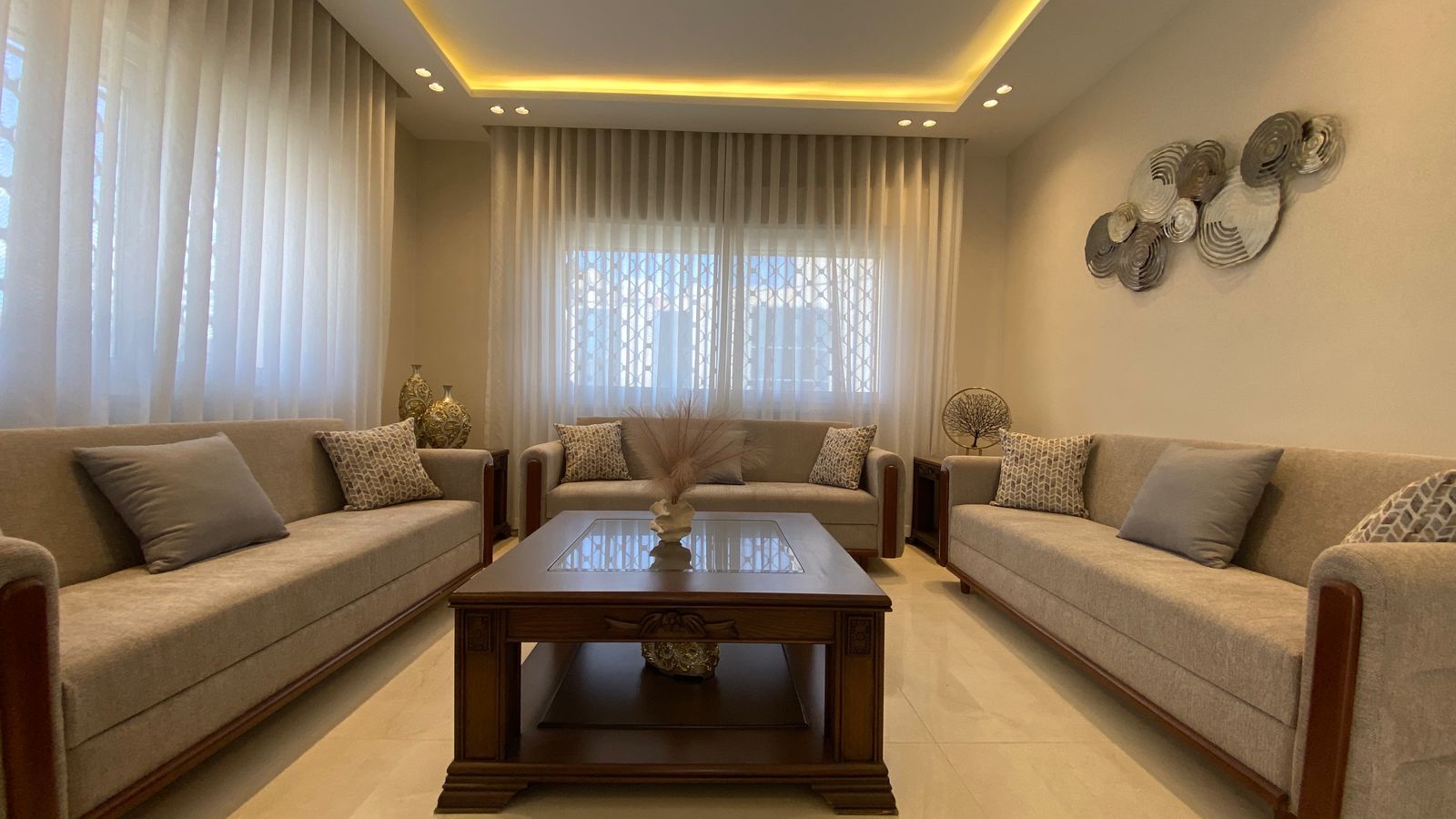 f4704ba3-7385-44e5-8901-cacd159ca943 Neutral living room with three beige sofas, gray throw pillows, and a wooden coffee table under warm ambient lighting.