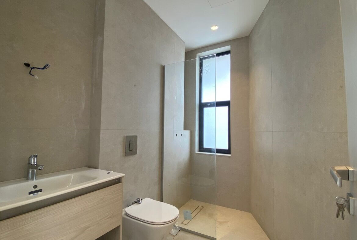 f475c0a7-7b03-4cc8-836e-0f6ea7d1efe1 Modern bathroom with beige large tiles, wall-mounted vanity and white rectangular sink, chrome faucet, and wall flush plate. A white toilet sits beside the vanity, and a frameless glass shower enclosure runs along the far wall with a tall narrow window above it. Recessed ceiling light and exposed electrical wires near the wall indicate ongoing finishing touches.
