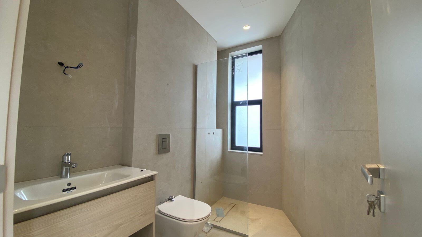 f475c0a7-7b03-4cc8-836e-0f6ea7d1efe1 Modern bathroom with beige large tiles, wall-mounted vanity and white rectangular sink, chrome faucet, and wall flush plate. A white toilet sits beside the vanity, and a frameless glass shower enclosure runs along the far wall with a tall narrow window above it. Recessed ceiling light and exposed electrical wires near the wall indicate ongoing finishing touches.