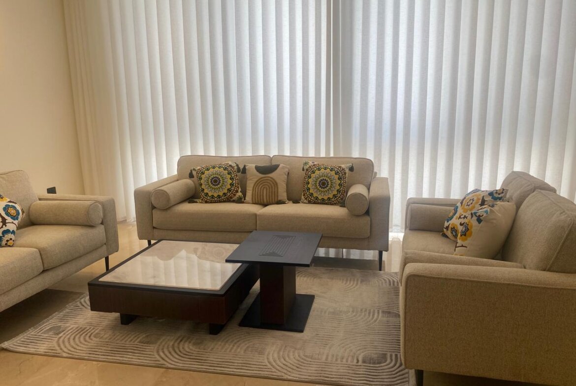 Living room with beige sofa set and decorative cushions, glass coffee table, and a light gray rug in front of white vertical blinds on windows.”,