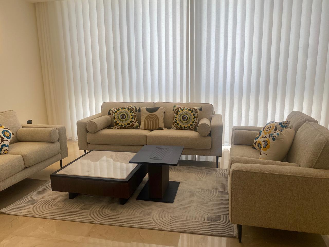 Living room with beige sofa set and decorative cushions, glass coffee table, and a light gray rug in front of white vertical blinds on windows.”,