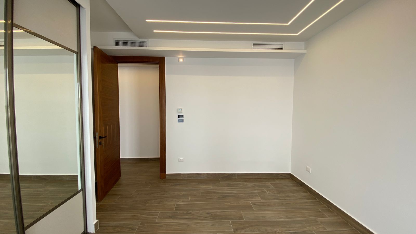 f48c868a-1aa1-43bb-a819-08ee83576dcd Empty modern room with wood door ajar, glass partition on left, and white walls; wood-look tile floor and geometric recessed ceiling lights.