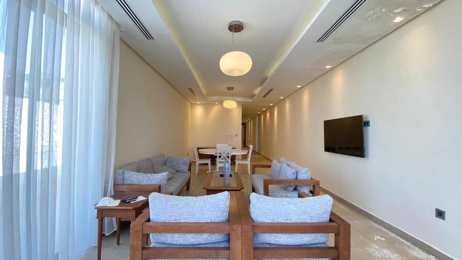 f49c70d9-db71-4b14-8933-f064fd8bc4e3 Open-plan living room with gray cushioned wooden-framed chairs around a glass-topped coffee table, and a wall-mounted TV on the right.