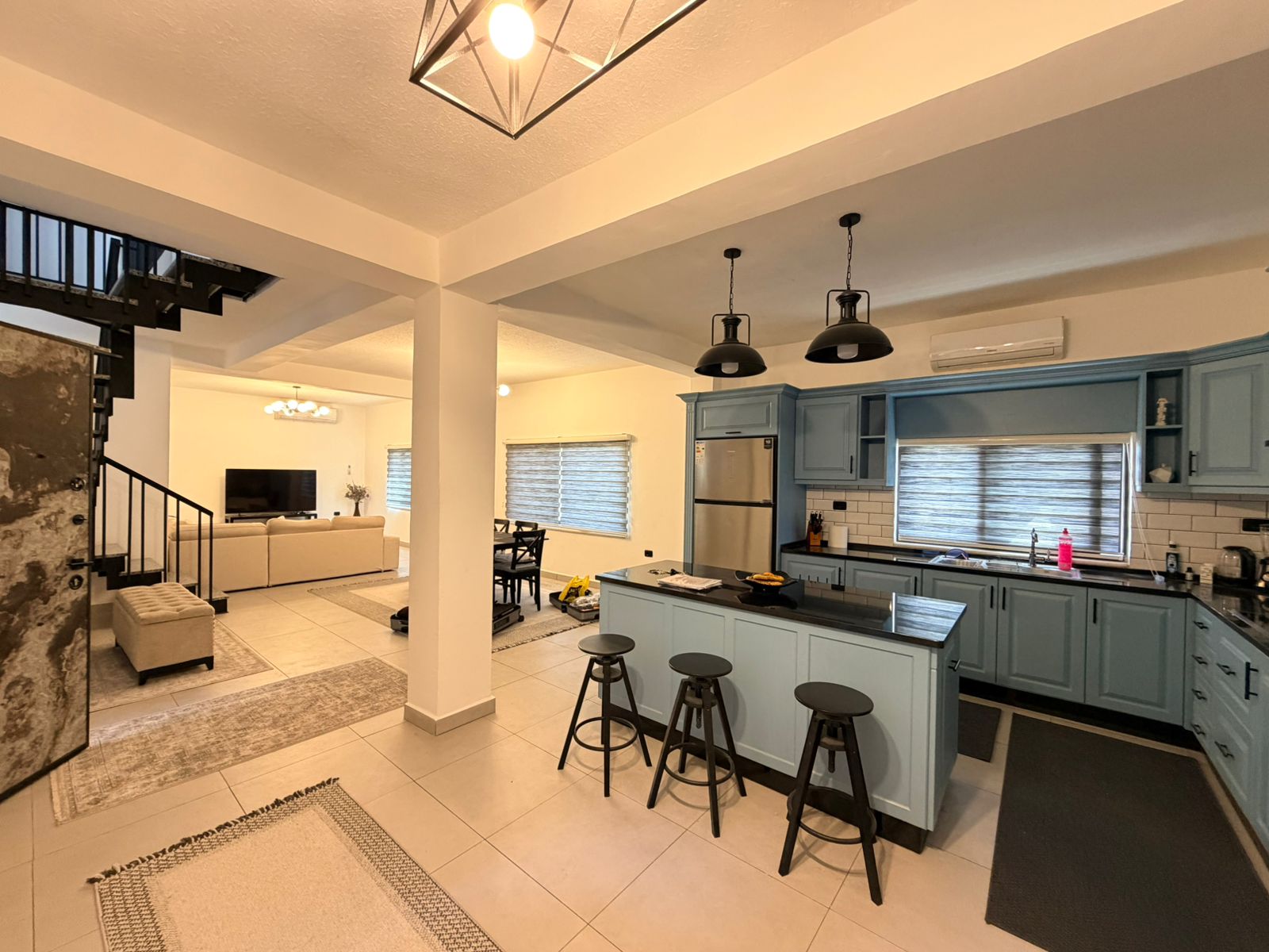 f4f104e4-d13d-4ad4-a84d-4eb9d324035d Open-concept kitchen with blue cabinets and a central island with three black bar stools, connected to a living room with a beige sofa and a TV area; staircase visible on the left, large windows with blinds, and tiled floors.