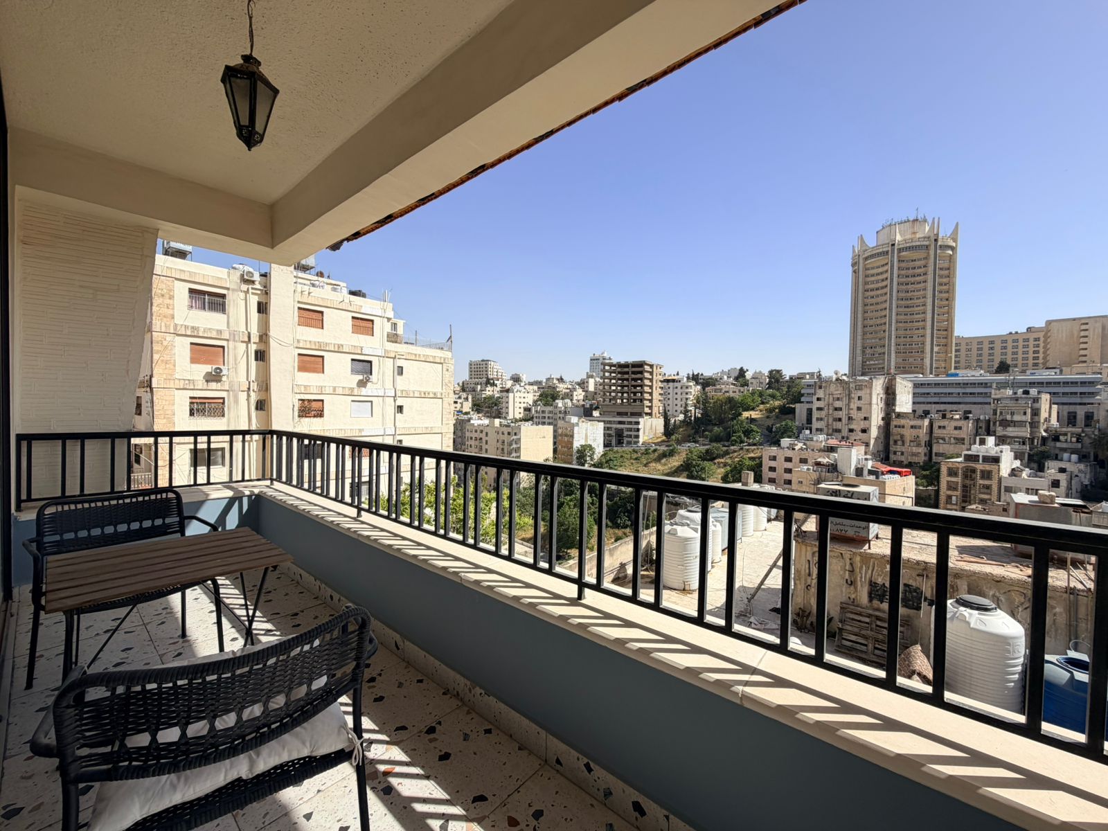 f5775063-d27d-486e-9079-49f16809991f Balcony seating area with wicker chairs and a small table, overlooking a dense city of beige buildings on a sunny day.