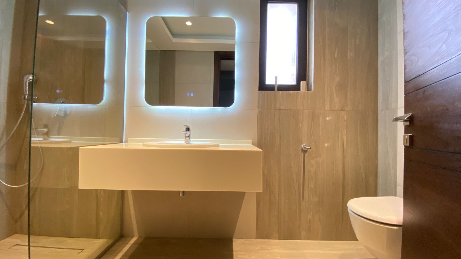 f5da06b2-b76c-4177-93ae-b34c6198ce27 Modern bathroom with backlit rectangular mirrors, floating sink, glass shower on the left, and a wall-mounted toilet on the right.