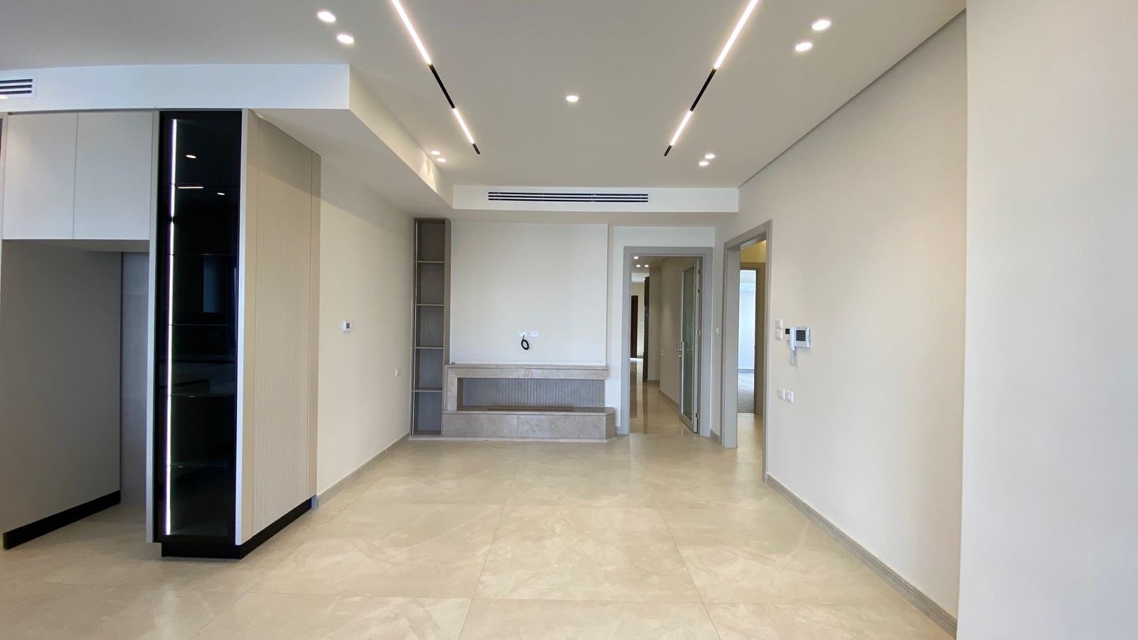 f5edd0f2-0dd4-4bad-99b4-0e192466be01 Empty modern living area with tiled beige floor, white walls, and built-in cabinets on the left; a marble reception-like counter at the far wall and a hallway opening to rooms on the right.