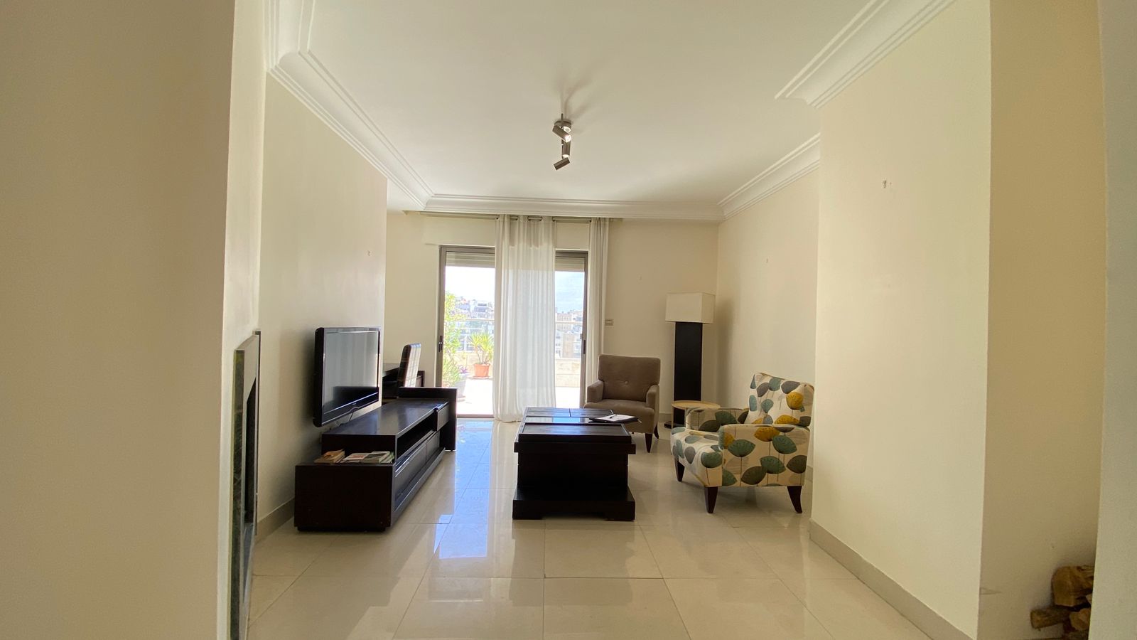 f644084a-e107-4a58-a07a-5adf58f72a77 Bright living room with white walls and glossy tile floor, featuring a dark wood coffee table, a wall-mounted TV and entertainment unit, two patterned armchairs and a beige chair, and sliding glass doors with sheer curtains opening to a balcony.